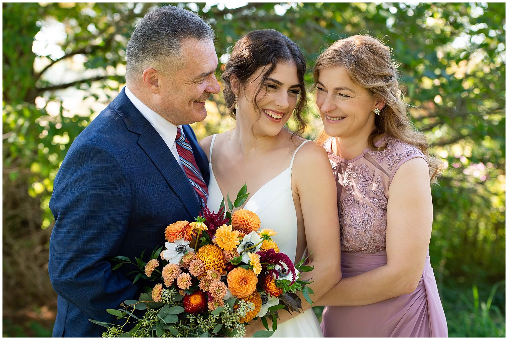 a bride with her mother and father. The bride is holding a fall coloured bouquet and they're laughing.