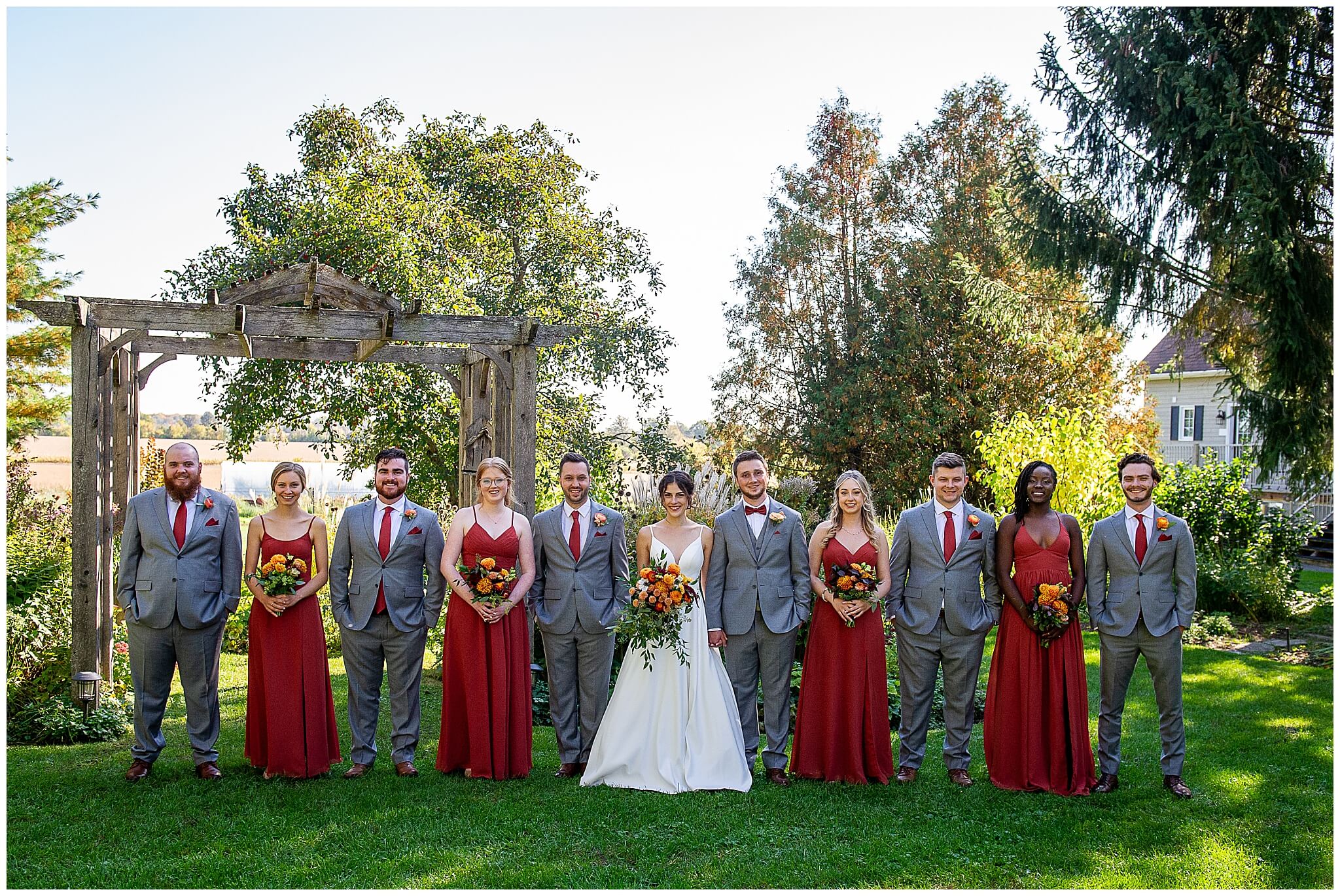 bridesmaids in long cinnamon coloured gowns holding fall bouquets and standing beside groomsmen with grey suits . Taken outdoors in the gardens of Strathmere Wedding venue near Ottawa