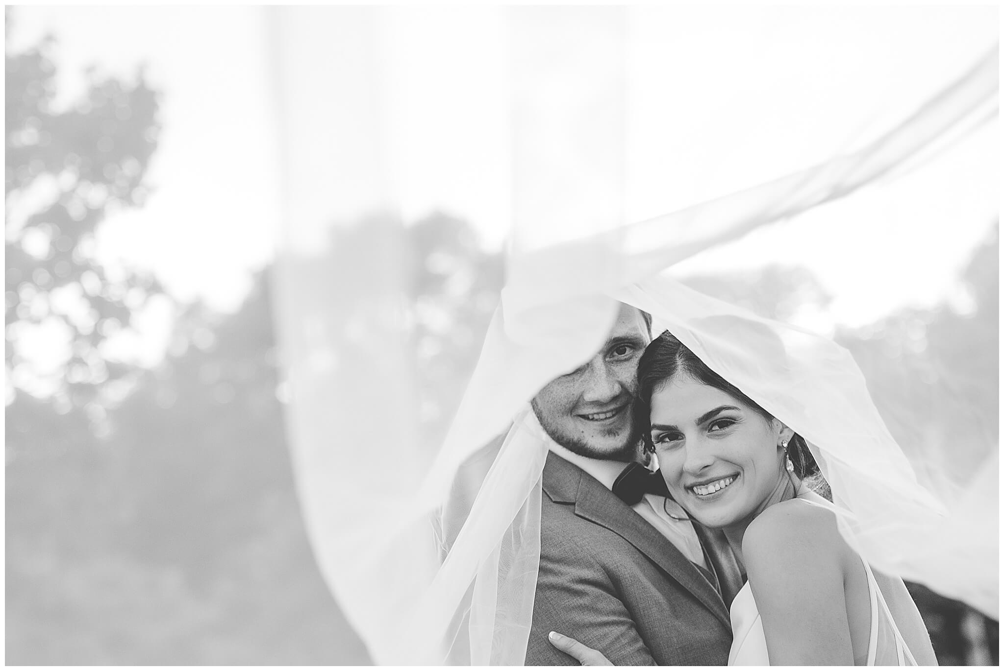 a playful black and white photo of a smiling bride and groom under a veil