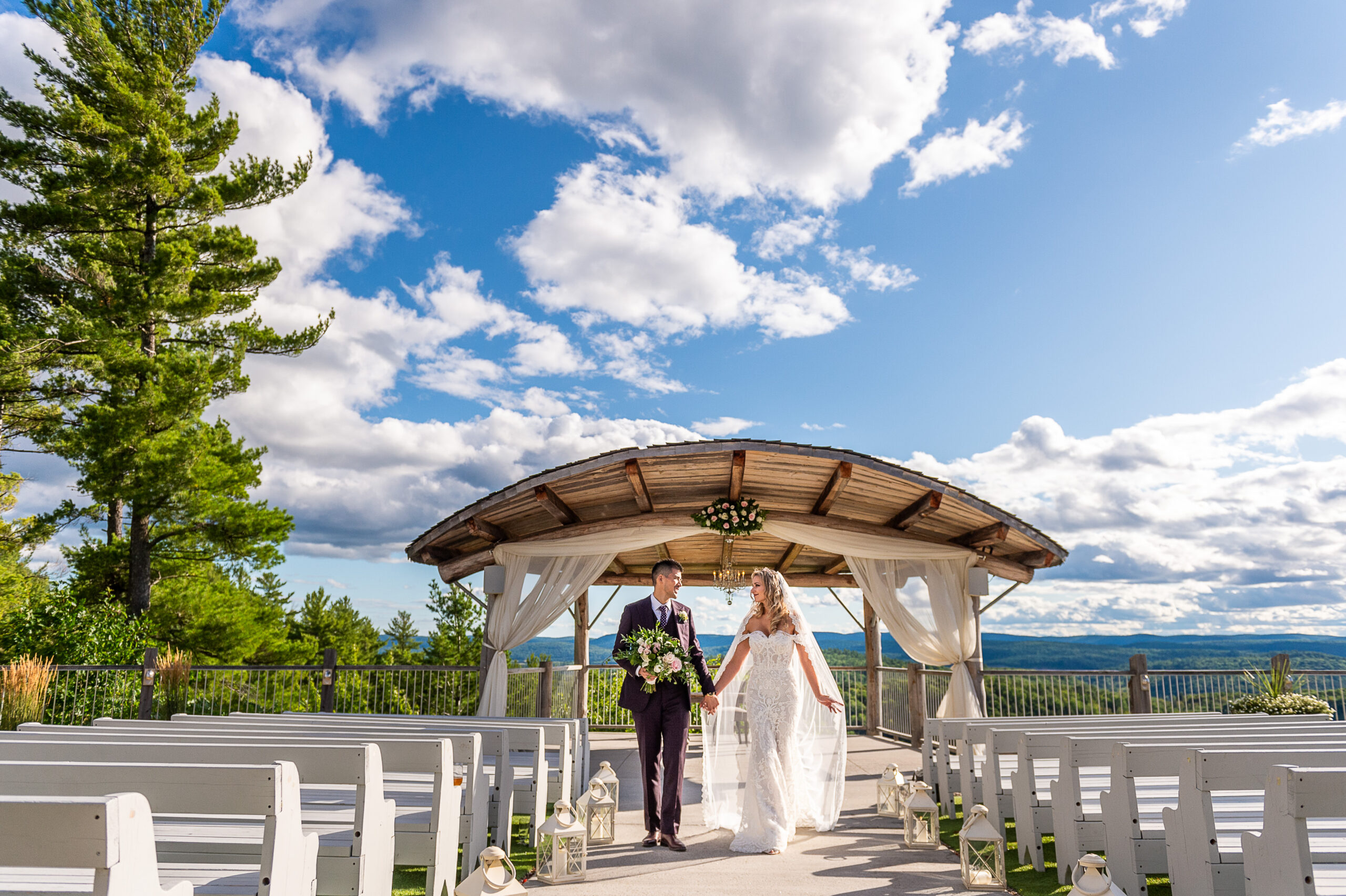 bride and groom walking hand in hand at their Le Belvedere ceremony site with a big blue sky around them. Captured outdoors by Ottawa Wedding Photographer JEMMAN Photography