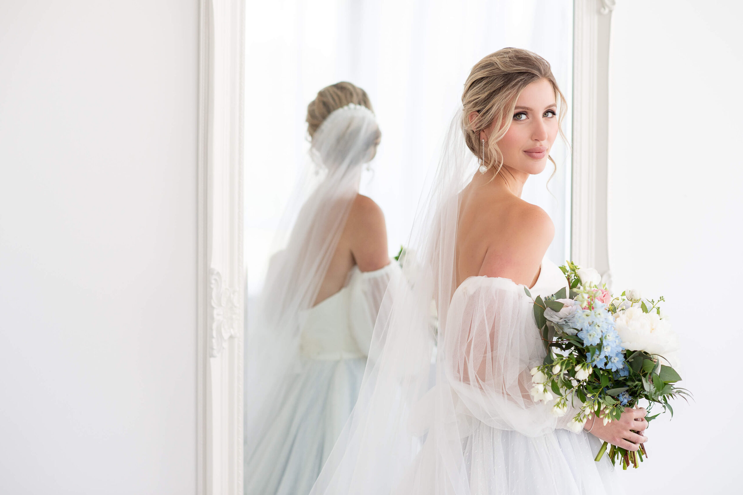 a bride wearing a veil and holding a bouquet of pink, white and blue flowers looking back over her shoulder with a reflection in the mirror. The happiness on the bride's face shows how to choose a wedding photographer in Ottawa