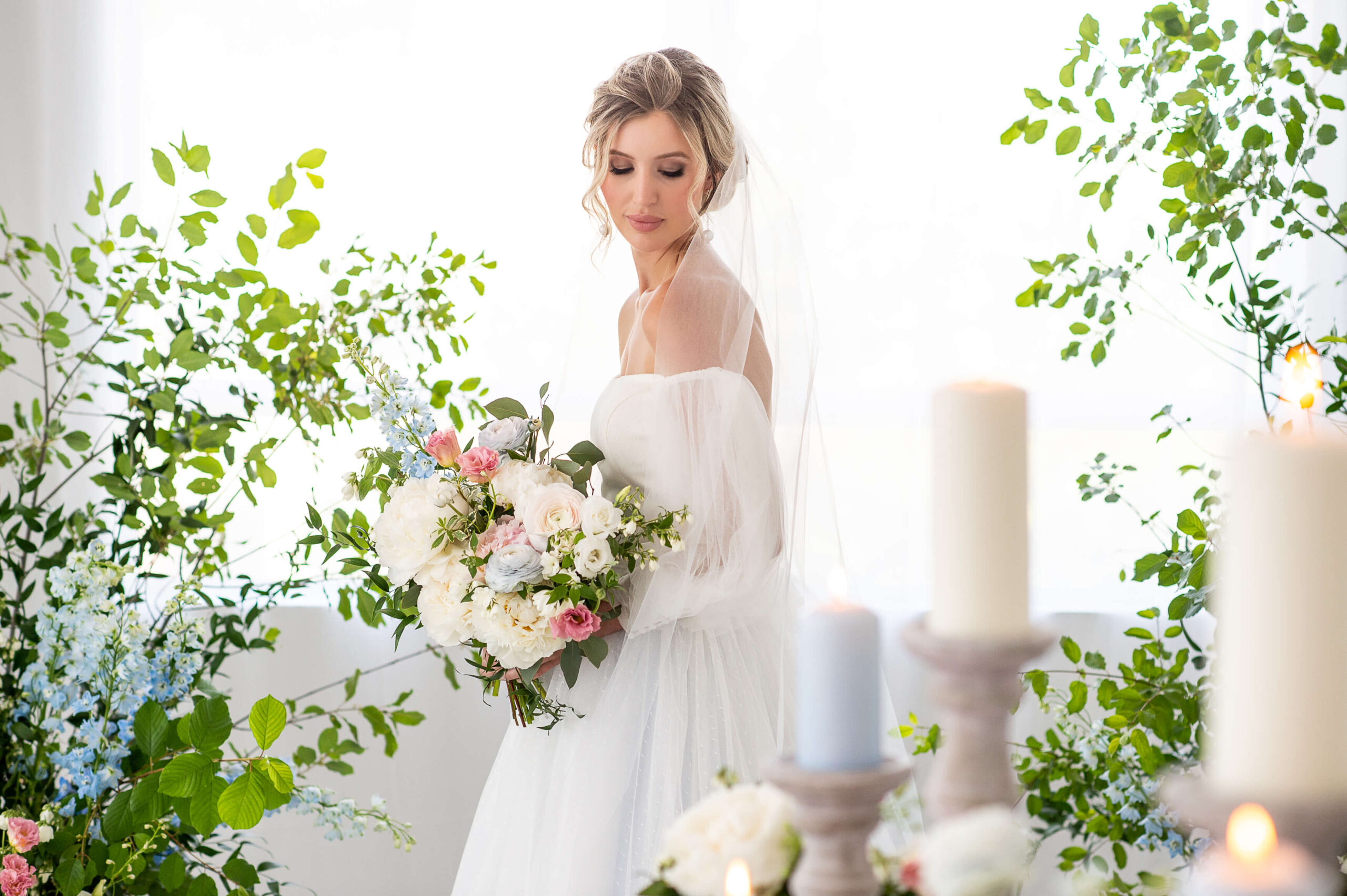 a photo of a bride looking down at her pink, blue and white bouquet. There are out of focus candles and greenery. Captured by Ottawa Wedding Photographer JEMMAN Photography