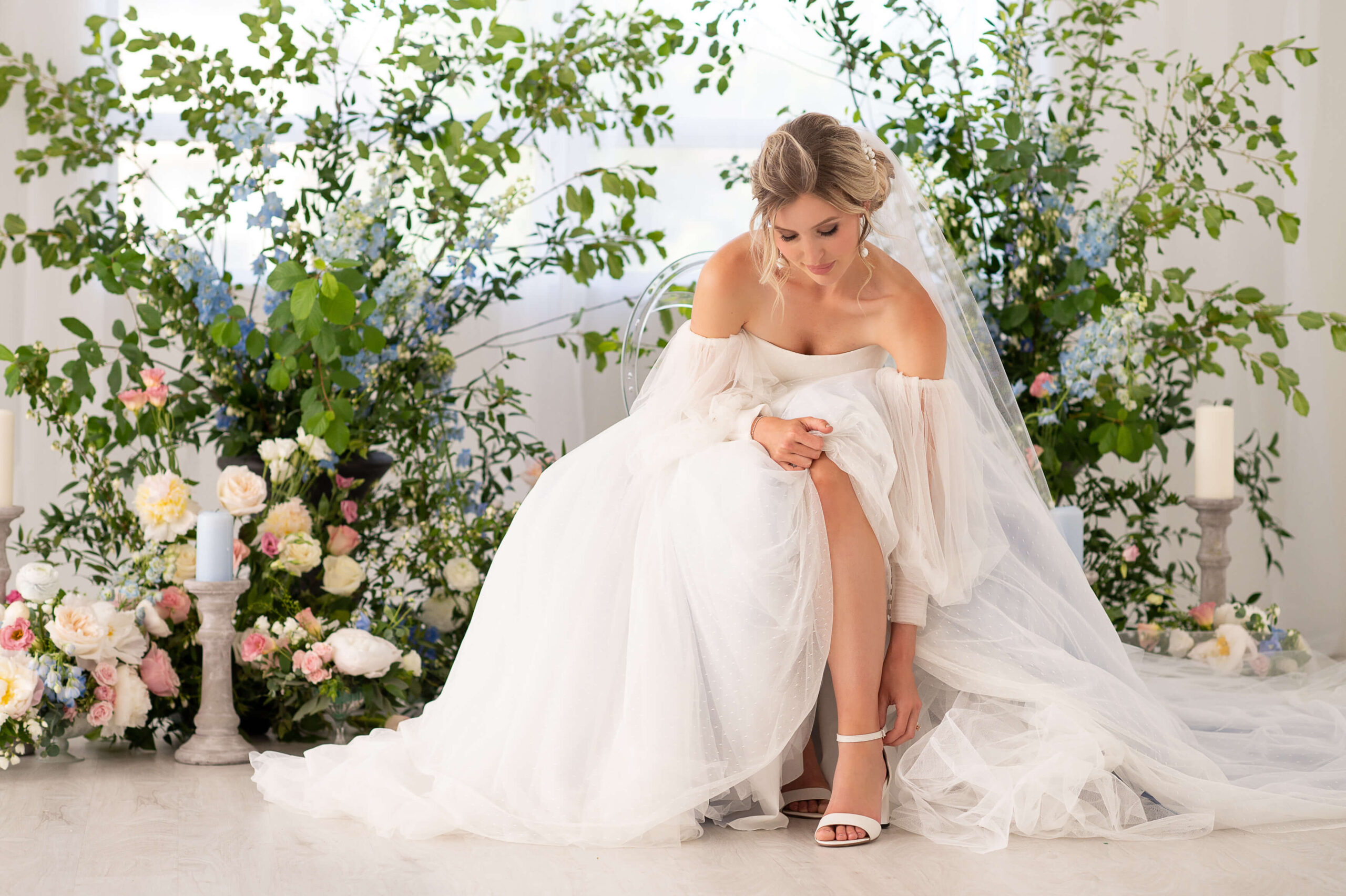 a bride sitting down adjusting her shoe in front of a wall of flowers. Capture indoors by Ottawa Wedding Photographer JEMMAN Photography