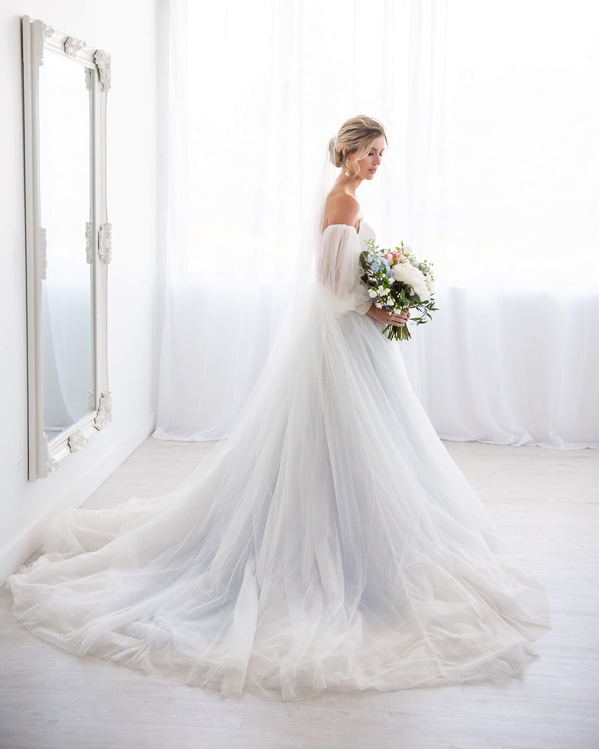 a full length portrait of a bride standing in front of a mirror holding her bouquet of pink, white and blue flowers. Captured by Ottawa Wedding Photographer JEMMAN Photography