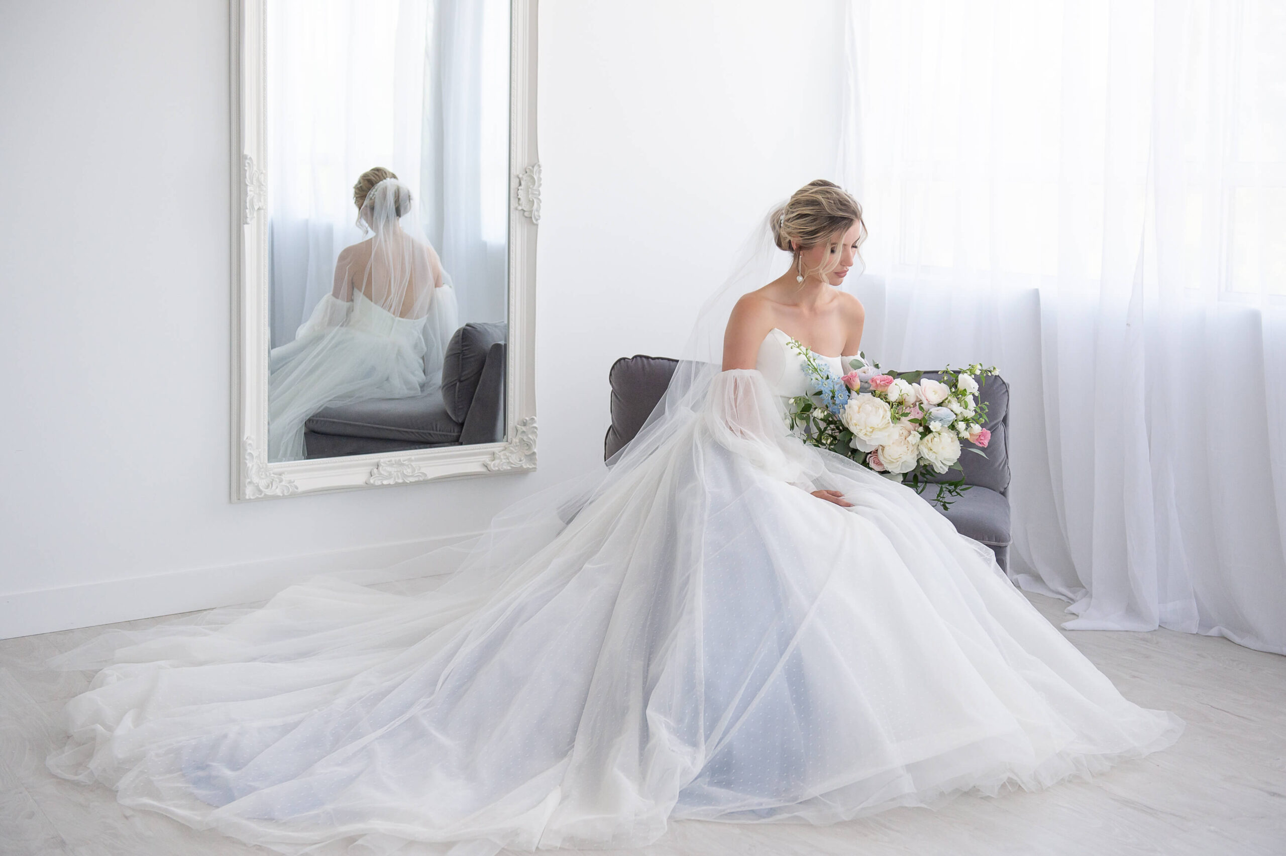a bride sitting on a grey couch holding a bouquet of blue, white and pink flowers. Image demonstrates the importance of how to choose a wedding photographer in Ottawa