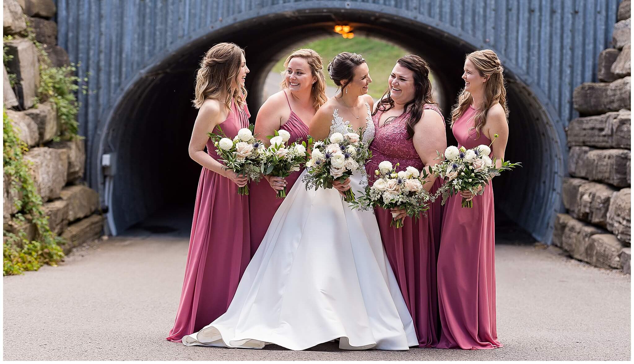 a bride and her bridesmaids in rose coloured dresses holding bouquets. Taken outside at The Marshes wedding venue