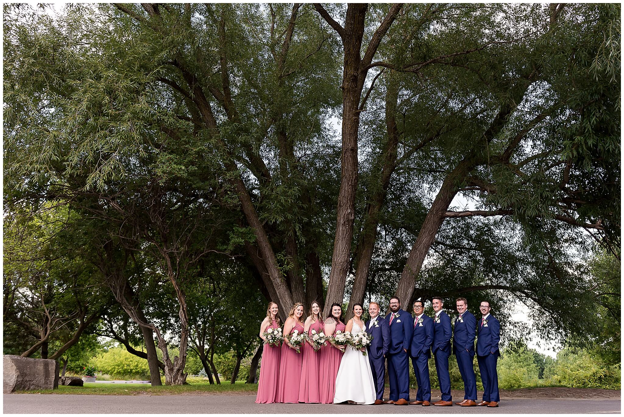 a group bridal party photo of bridesmaids in long rose dresses and groomsmen in blue suits underneath the large trees at The Marshes wedding venue