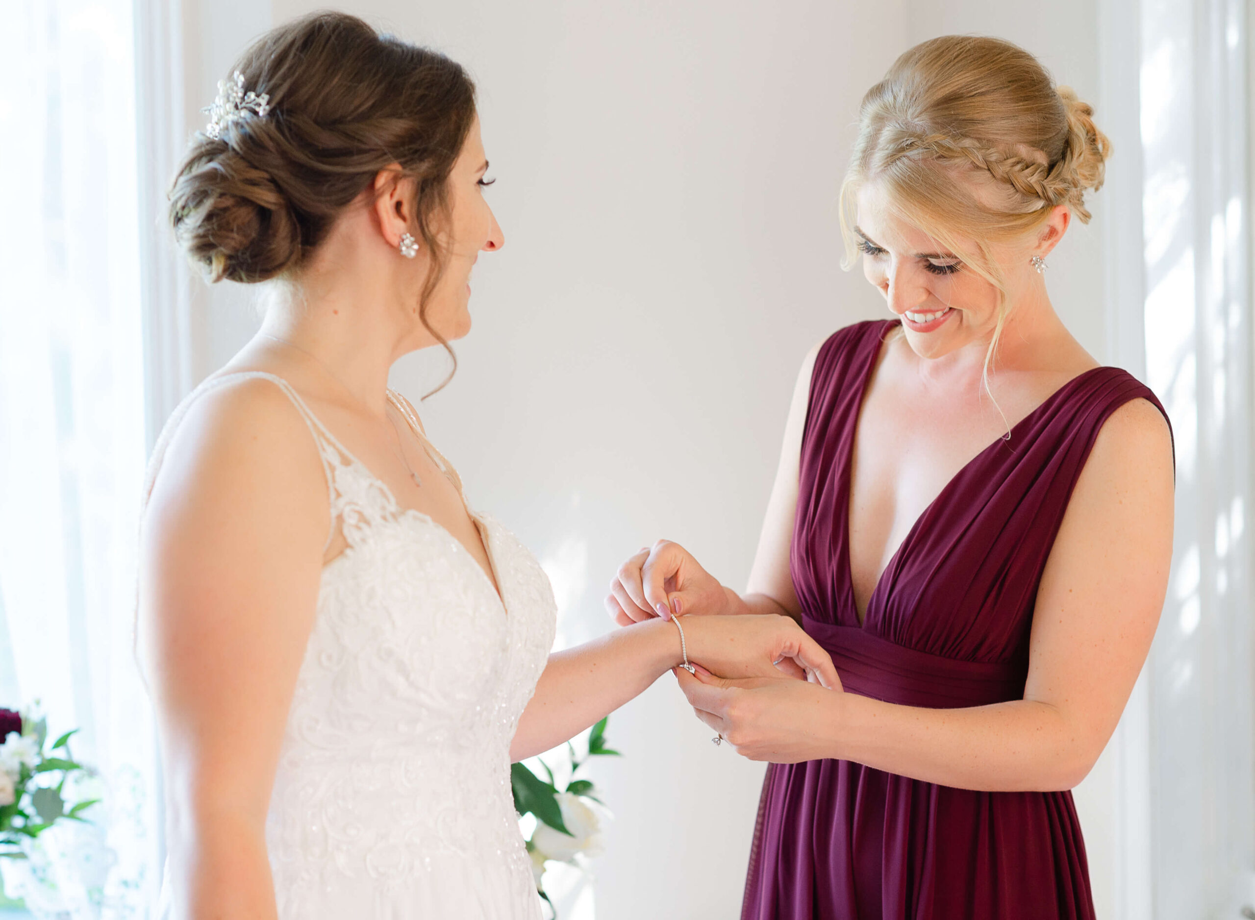 a bridesmaid in a burgundy gown places a bracelet on a bride's wrist