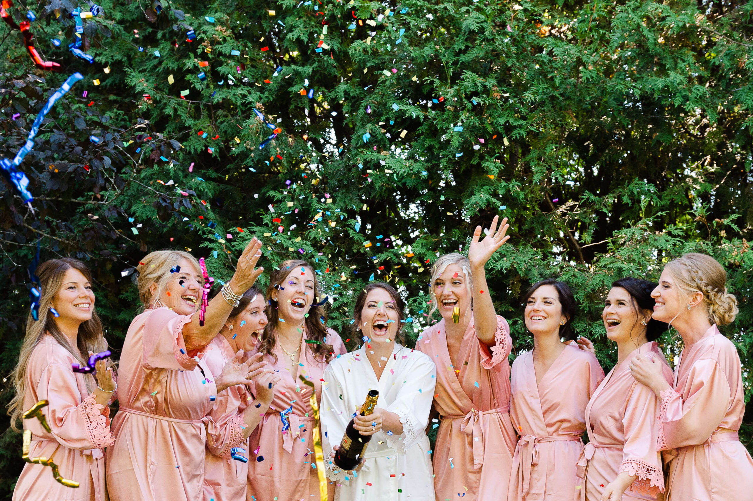 a bride popping a champagne streamer bottle with her bridesmaids dressed in pink robes. Wedding organized by Opportunity Knocks Events