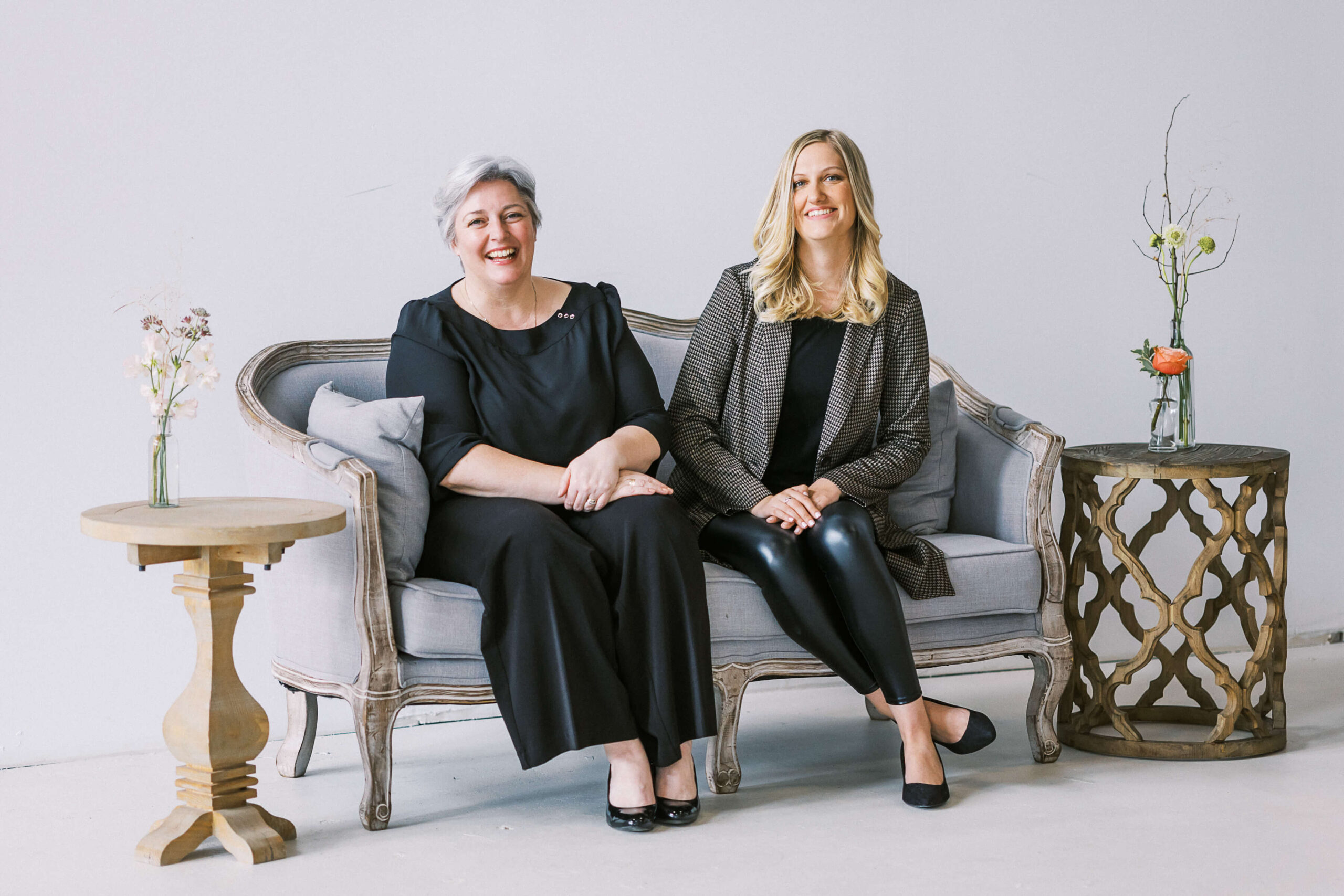Kiley and Janet from Opportunity Knocks Events sitting on a grey settee, dressed in black laughing and smiling