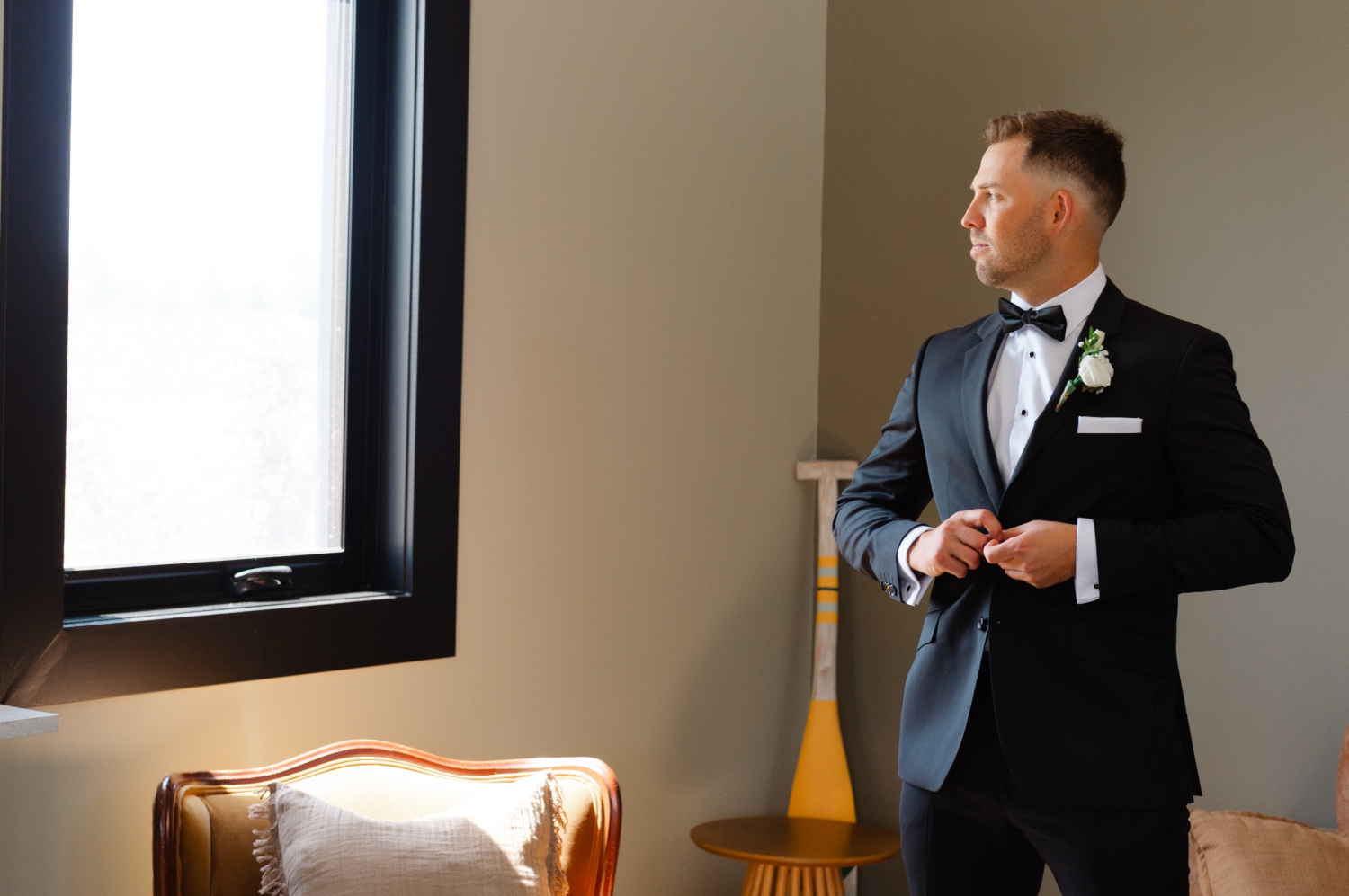 a groom buttons his jacket as he looks out the window of the groom's suite at the Bleeks and Bergamot Wedding Venue