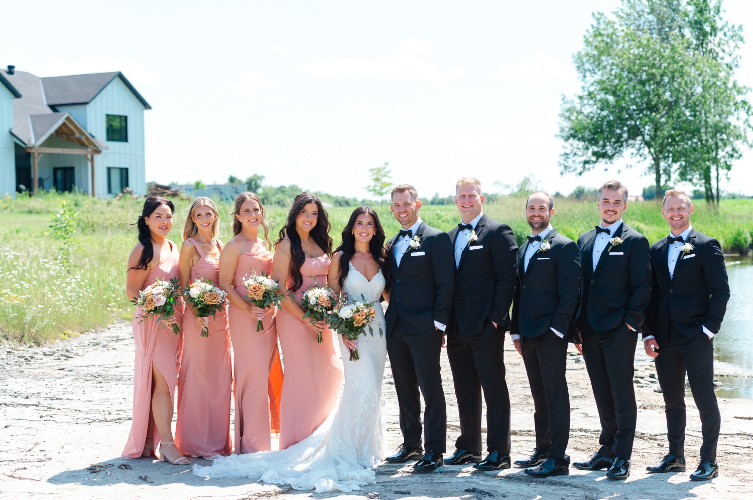 a formal portrait of a bride and groom and their bridesmaids in pink gowns and their groomsmen in tuxes on the riverside at the Bleeks and Bergamot Wedding Venue