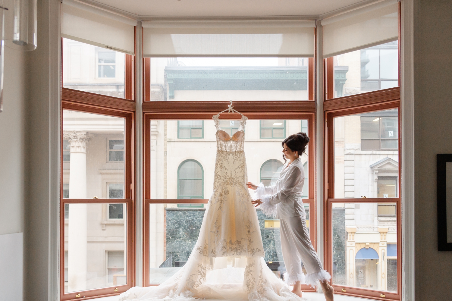 a bride in white pajamas touching her gown as it hangs in the window of the Restays hotel. This photo is part of her Museum of Nature Wedding Photos
