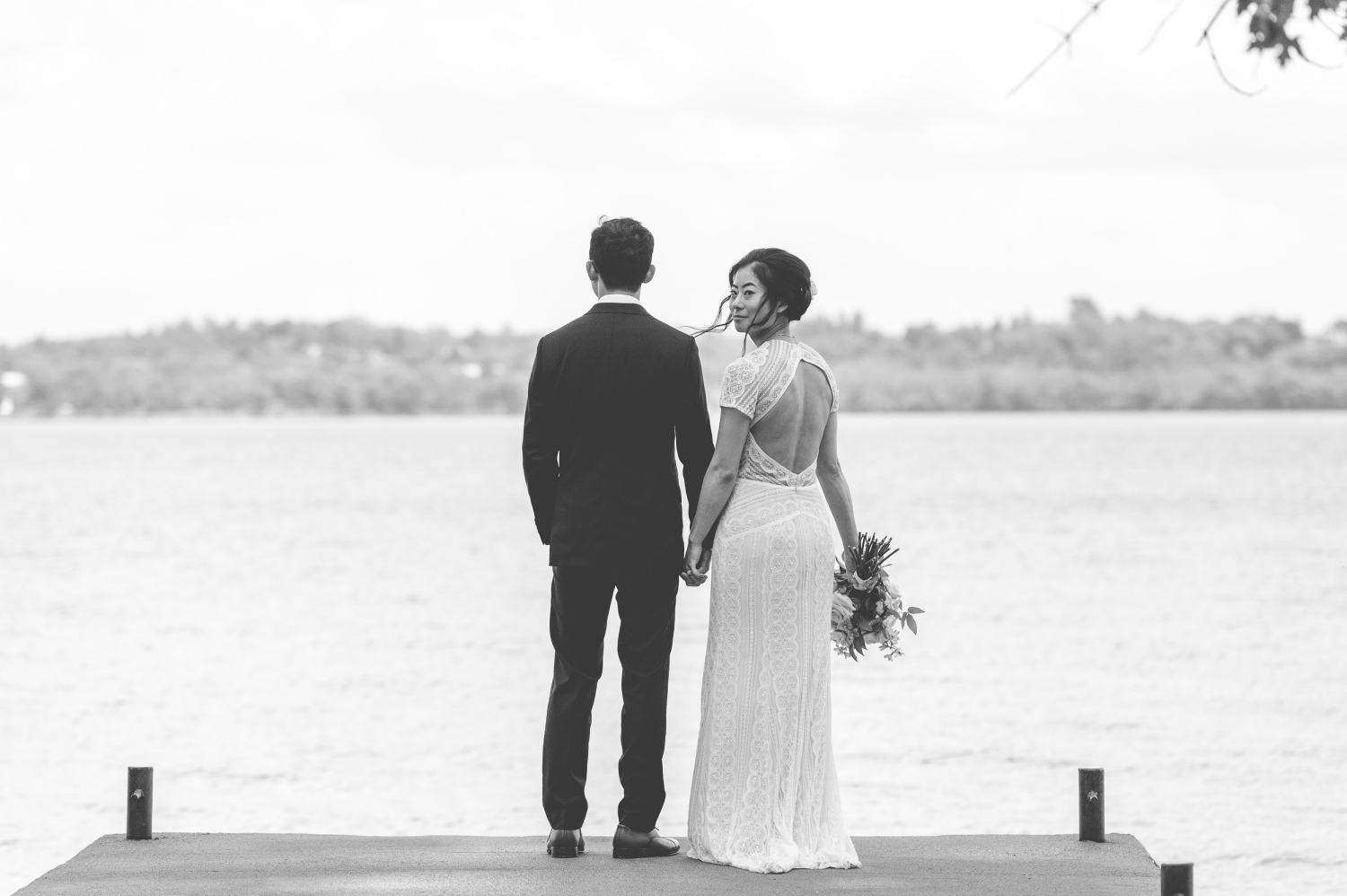 a black and white photo of a bride and groom standing at the end of the dock during their Ottawa backyard wedding. The bride is looking behind her