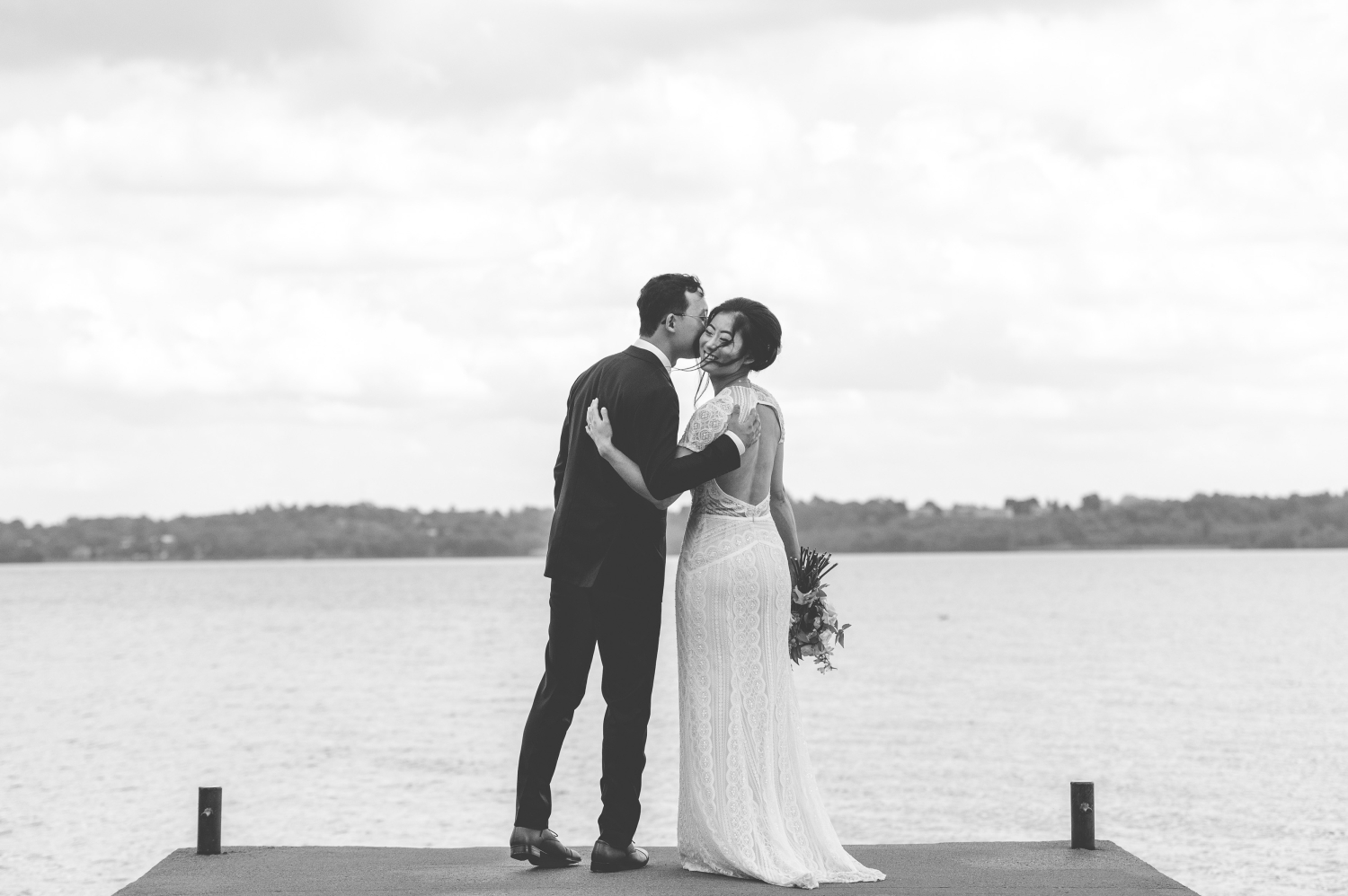 a black and white photo of a groom kissing his bride on a dock. Captured by JEMMAN Photography as part of their Ottawa backyard wedding photography