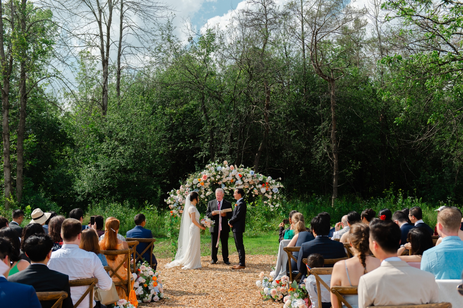 a bride and groom at the altar with a floral archway behind them and their guests looking on. Captured as part of an Ottawa backyard wedding