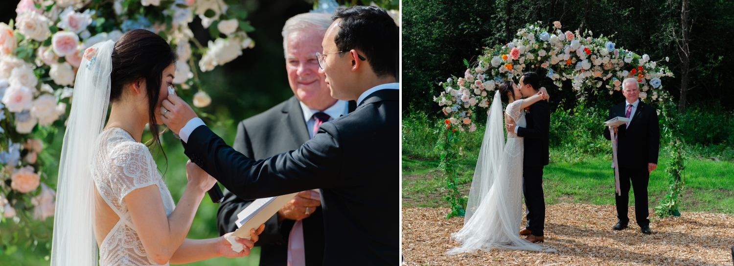 a groom wipes away his bride's tears and kisses her during their wedding ceremony. There is a floral arch and an officiant in the background