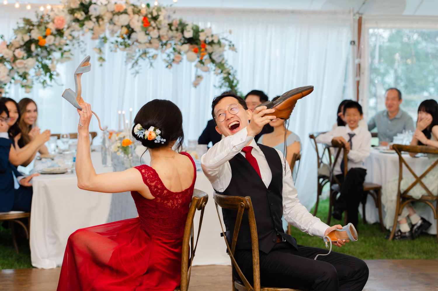 the shoe game where a bride in a red dress and a groom in a black suit hold up their shoes in response to questions. Captured by JEMMAN Photography during an Ottawa backyard wedding