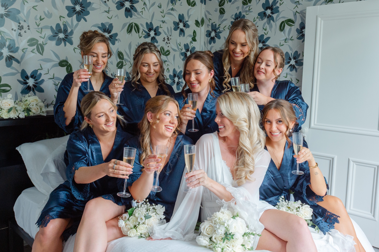 a group photo of a bride in a white robe and her bridesmaids in blue robes sitting on a bed and toasting with champagne. Captured by JEMMAN Photography at Maple and Rose Luxury Accommodations