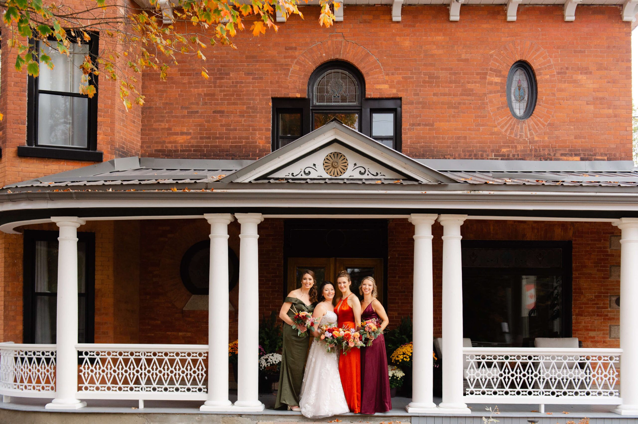 a bride with her bridesmaids in fall coloured dresses standing on the front porch of the Maple and Rose wedding venue and accommodations. Captured by JEMMAN Photography