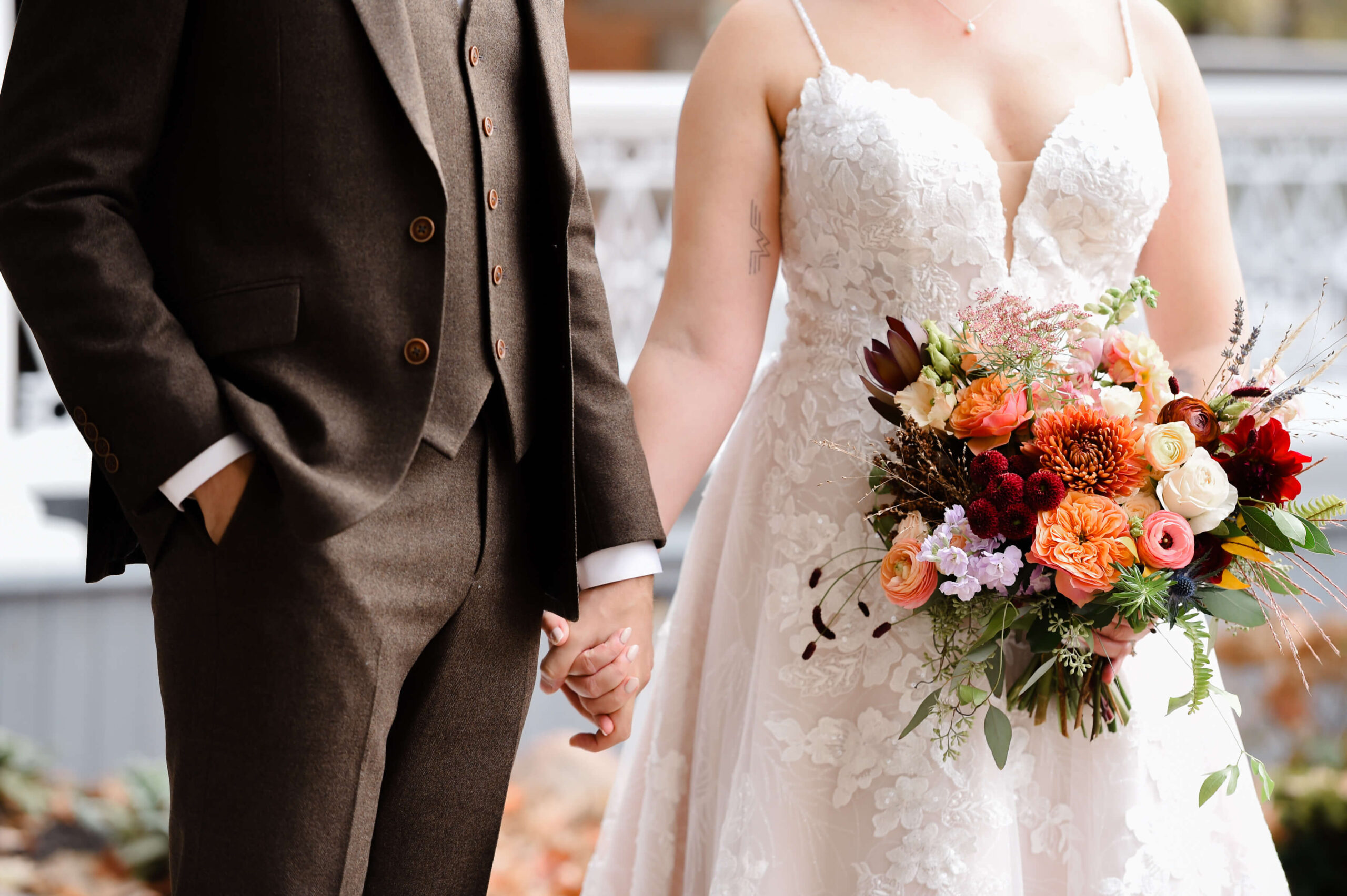 a closeup photo of a bride holding a fall coloured bouquet and holding the hand of her groom in a brown suit. Captured outdoors by Ottawa wedding photographer JEMMAN Photography