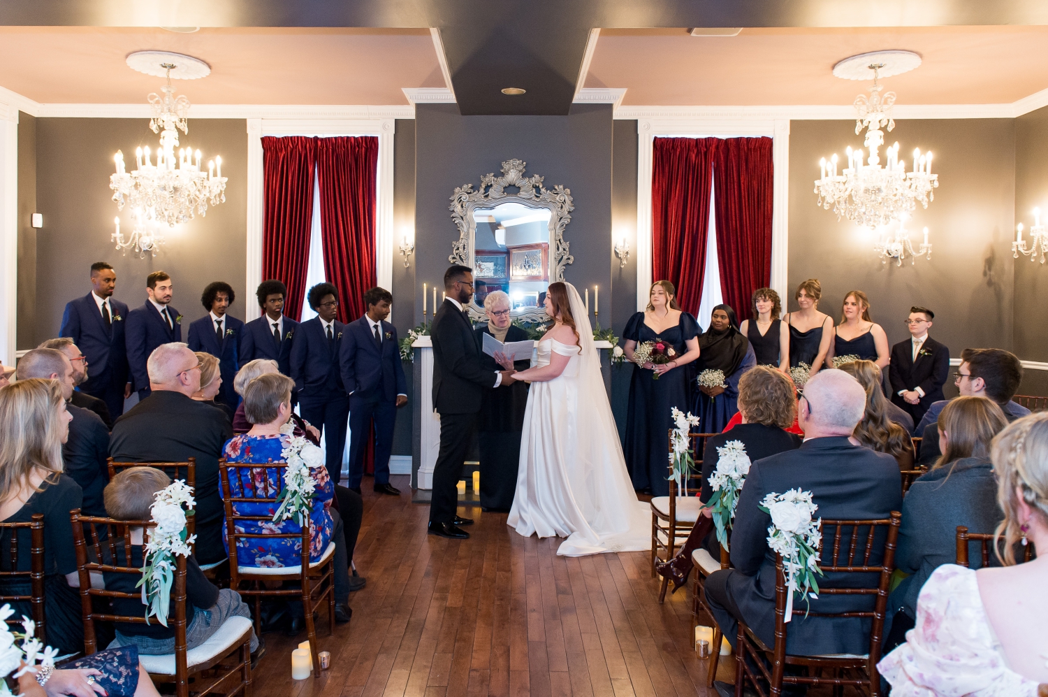 a bride and groom with their wedding party dressed in blue in front of the fireplace ceremony space of the Grand Hotel Weddings. Captured by JEMMAN Photography