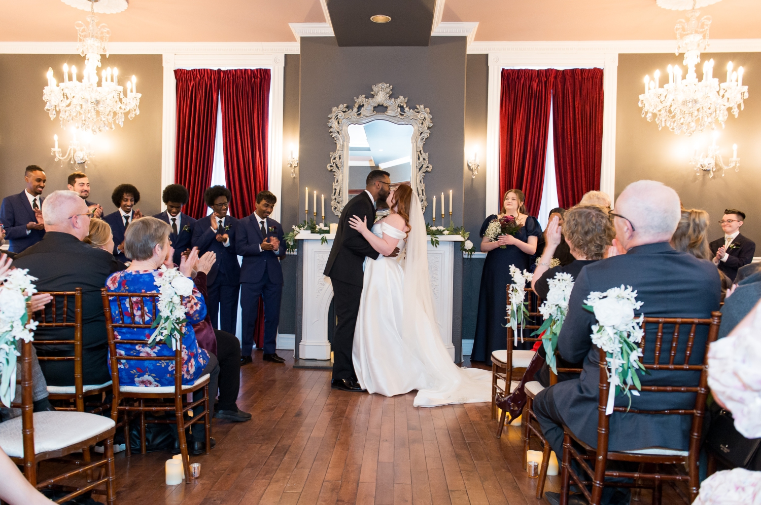 a bride and groom share a first kiss in the Grand Hotel wedding ceremony space surrounded by their clapping wedding party dressed in blue