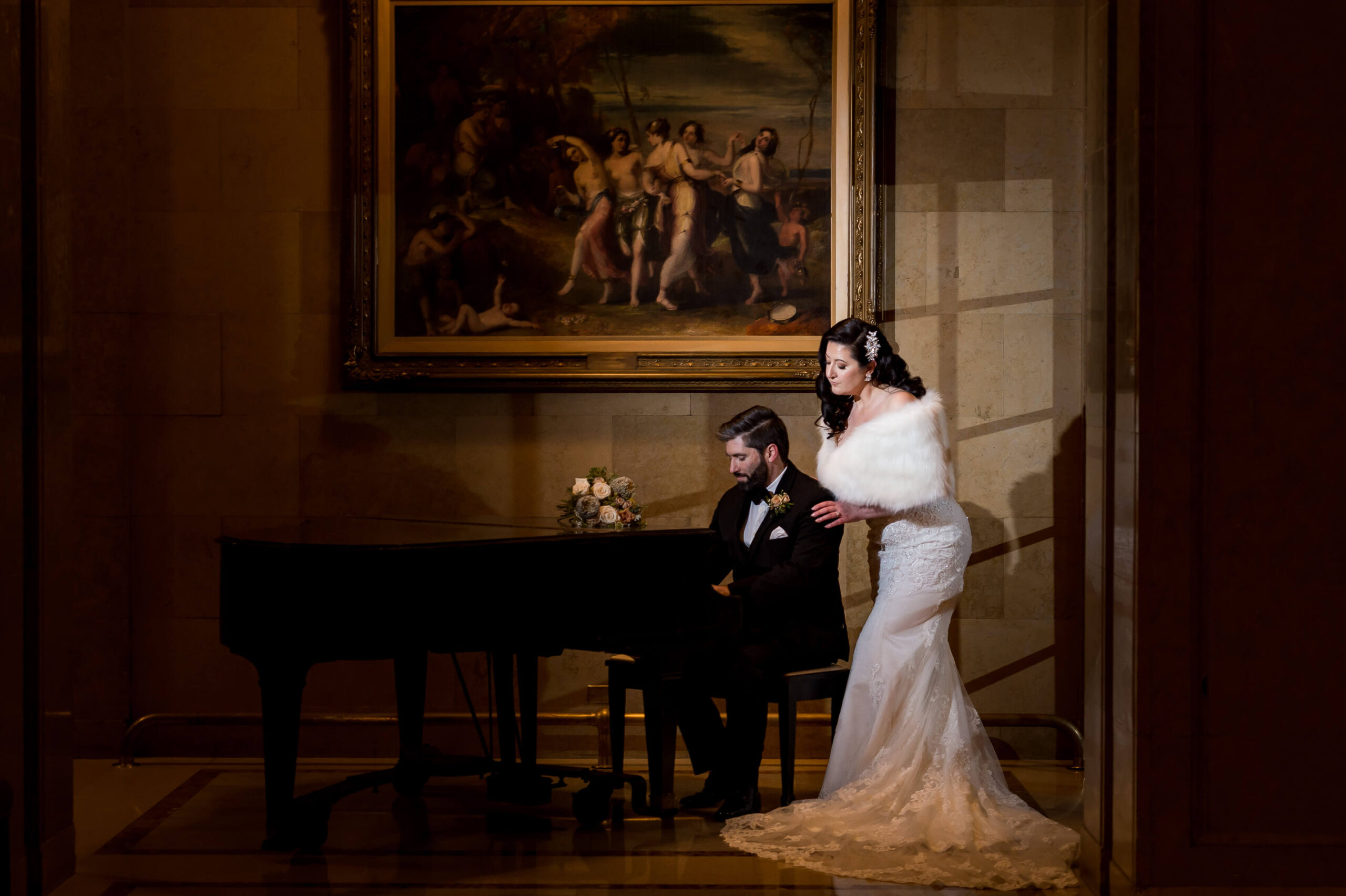 a photo of an elegant bride with fur stole looking over the shoulder of a groom in a black tuxedo playing on a grand piano in one of the grand hallways of the Chateau Laurier, a downtown Ottawa wedding venue. Captured by JEMMAN Photography