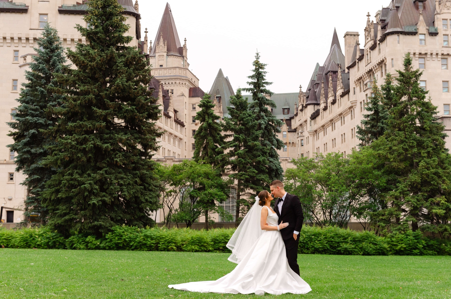 a sweeping photo of a bride in a white ballgown and a groom in a black tuxedo kissing behind the Chateau Laurier, a downtown Ottawa wedding venue. Captured outdoors by JEMMAN Photography