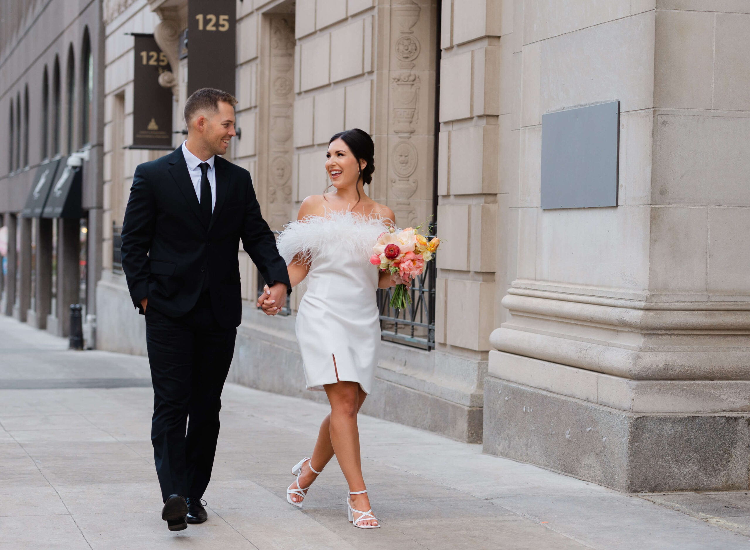 a bride in a short white dress and feathers walking and holding hands with a groom in a black suit outside the Ottawa Art Gallery. Captured by Ottawa wedding photographer JEMMAN Photography