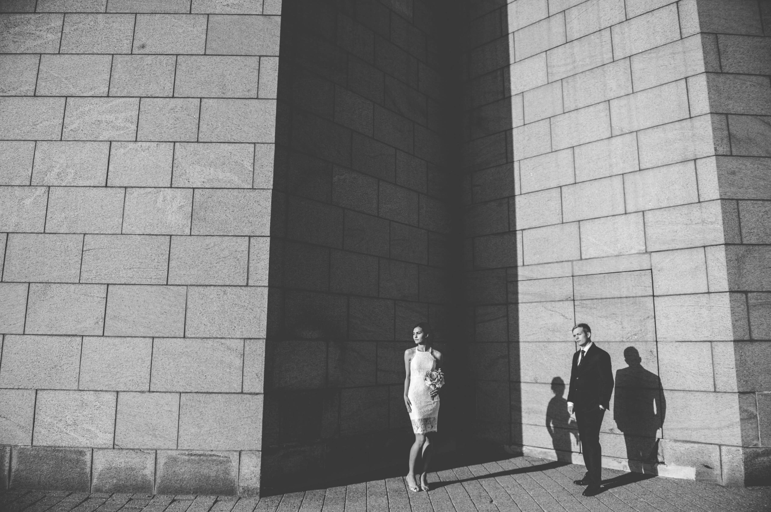 a black and white photo of a bride in a short white lace dress and a groom in a black suit standing in the sun and shade of the National Gallery of Canada. Captured by Ottawa wedding photographer JEMMAN Photography