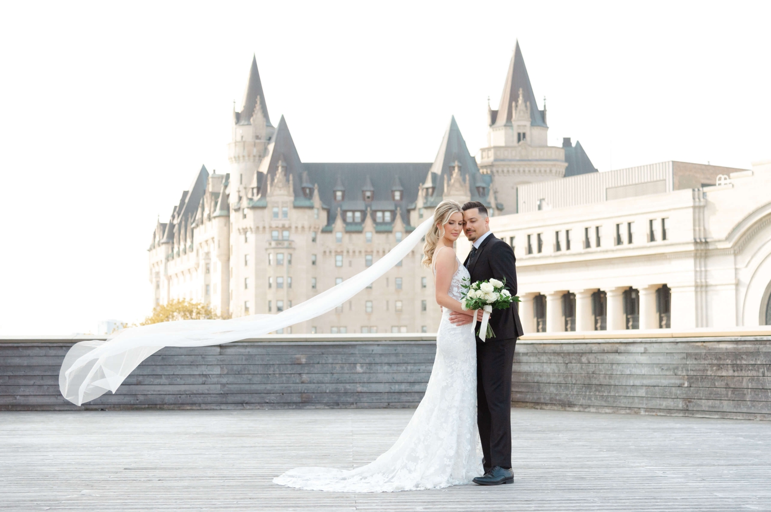 a groom in a black suit and a bride in a white lace dress with her veil flowing. Captured on the terrace of the NAC, a downtown Ottawa wedding venue, by JEMMAN Photography