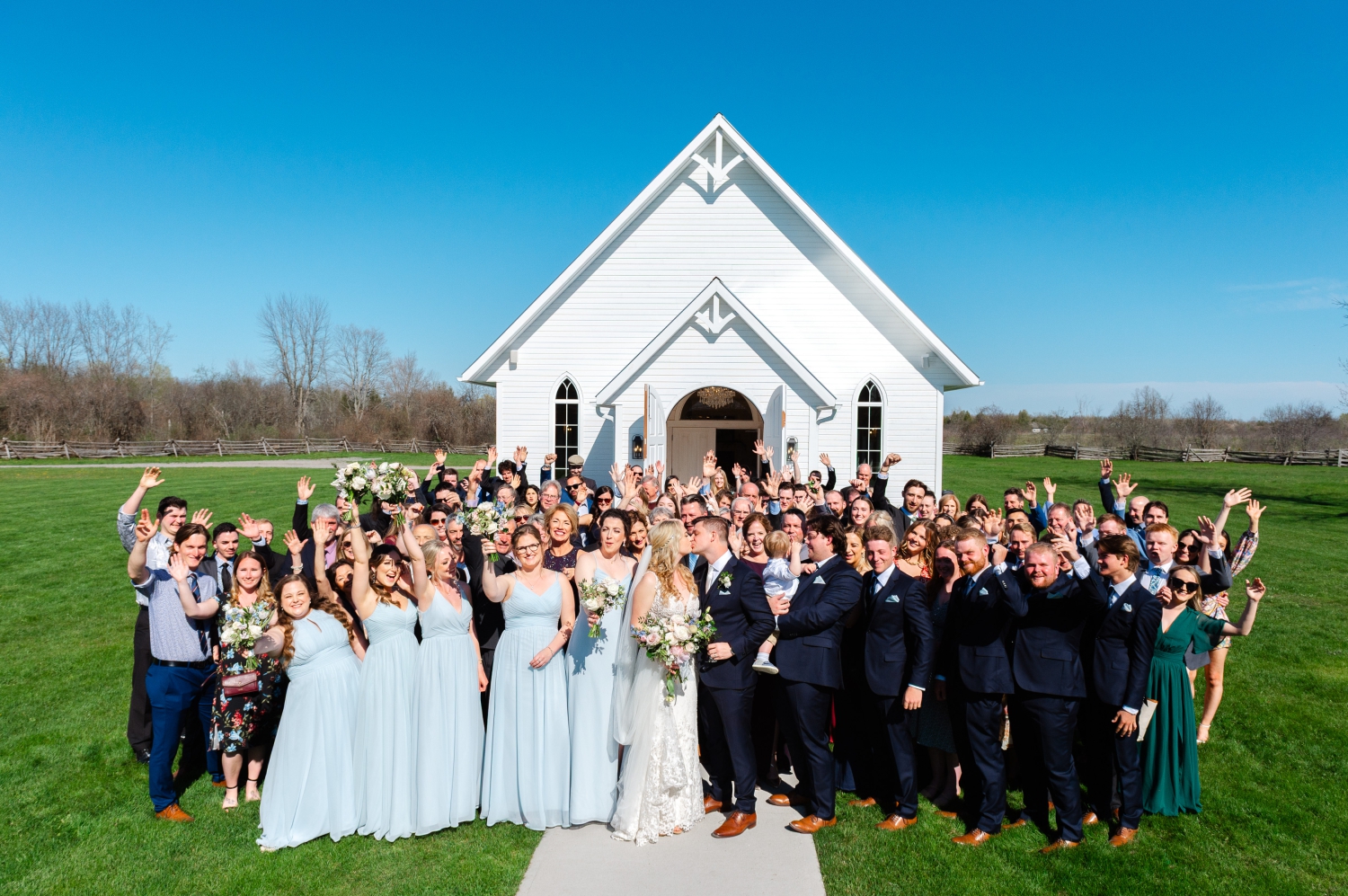 a group photo of a bride and groom kissing with all of their guests around them as they stand outside of the Ceremony House at Stonefields Estate, an Ottawa outdoor wedding venue. Captured by JEMMAN Photography