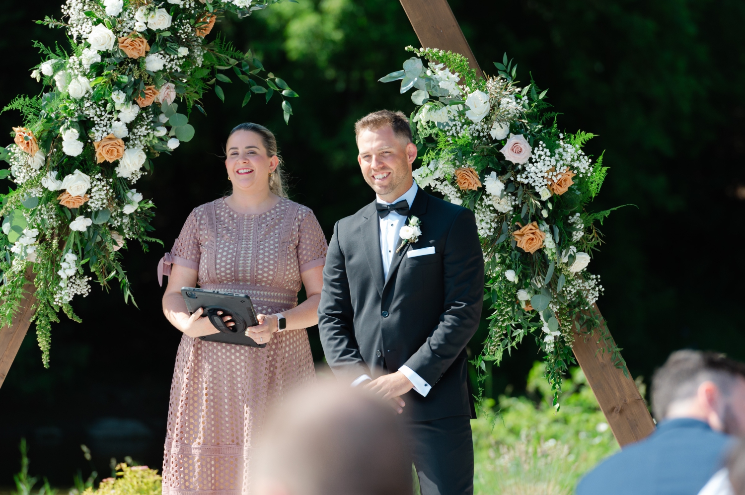 a groom in a black tux smiling as he watches his bride walk down the aisle of their Bleeks and Bergamot wedding. Captured by Ottawa wedding photographer JEMMAN Photography