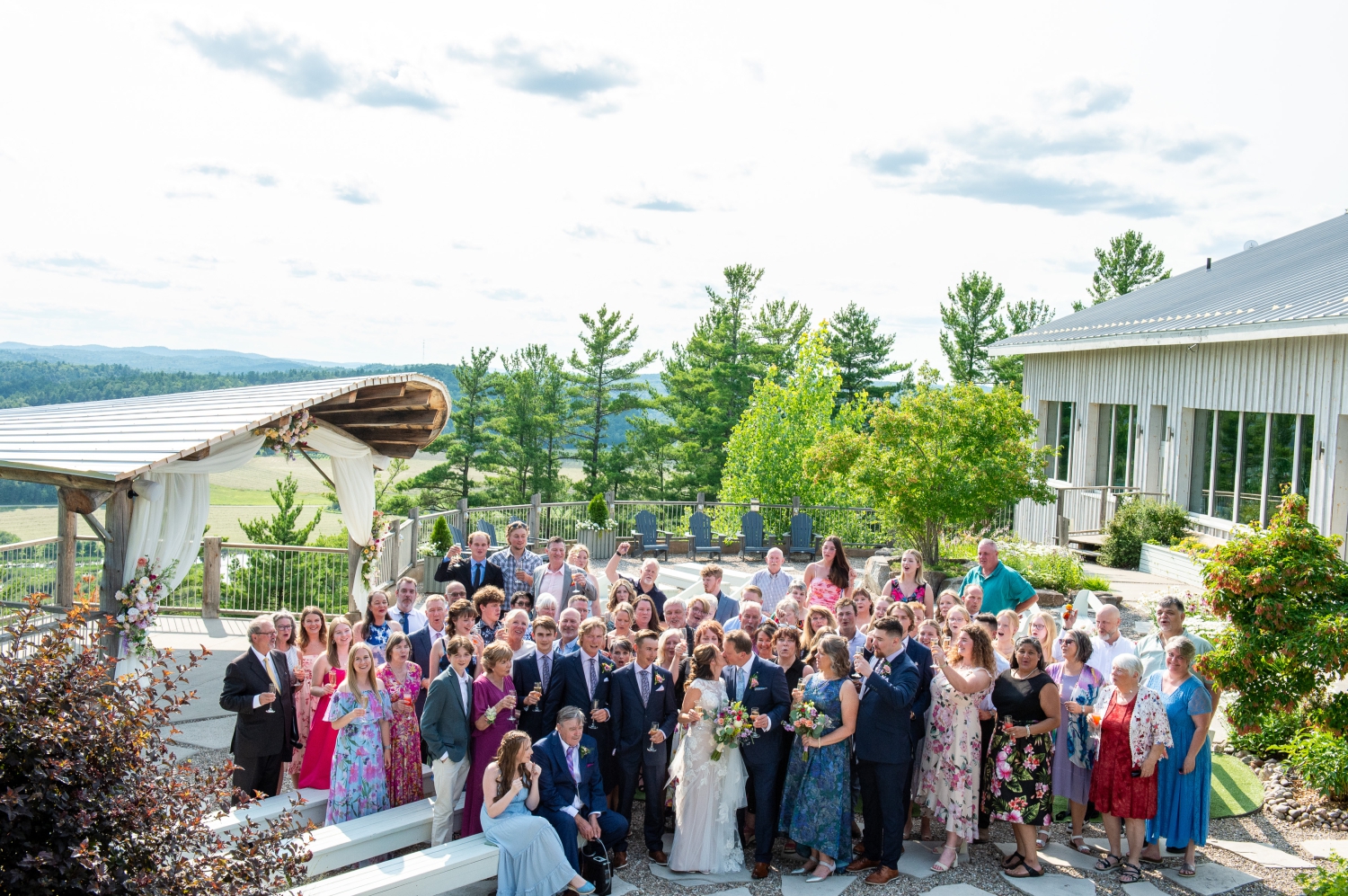 a group photo of a bride and groom kissing with all their guests behind them. Captured in the ceremony space of the Ottawa outdoor wedding venue, Le Belvedere by JEMMAN Photography