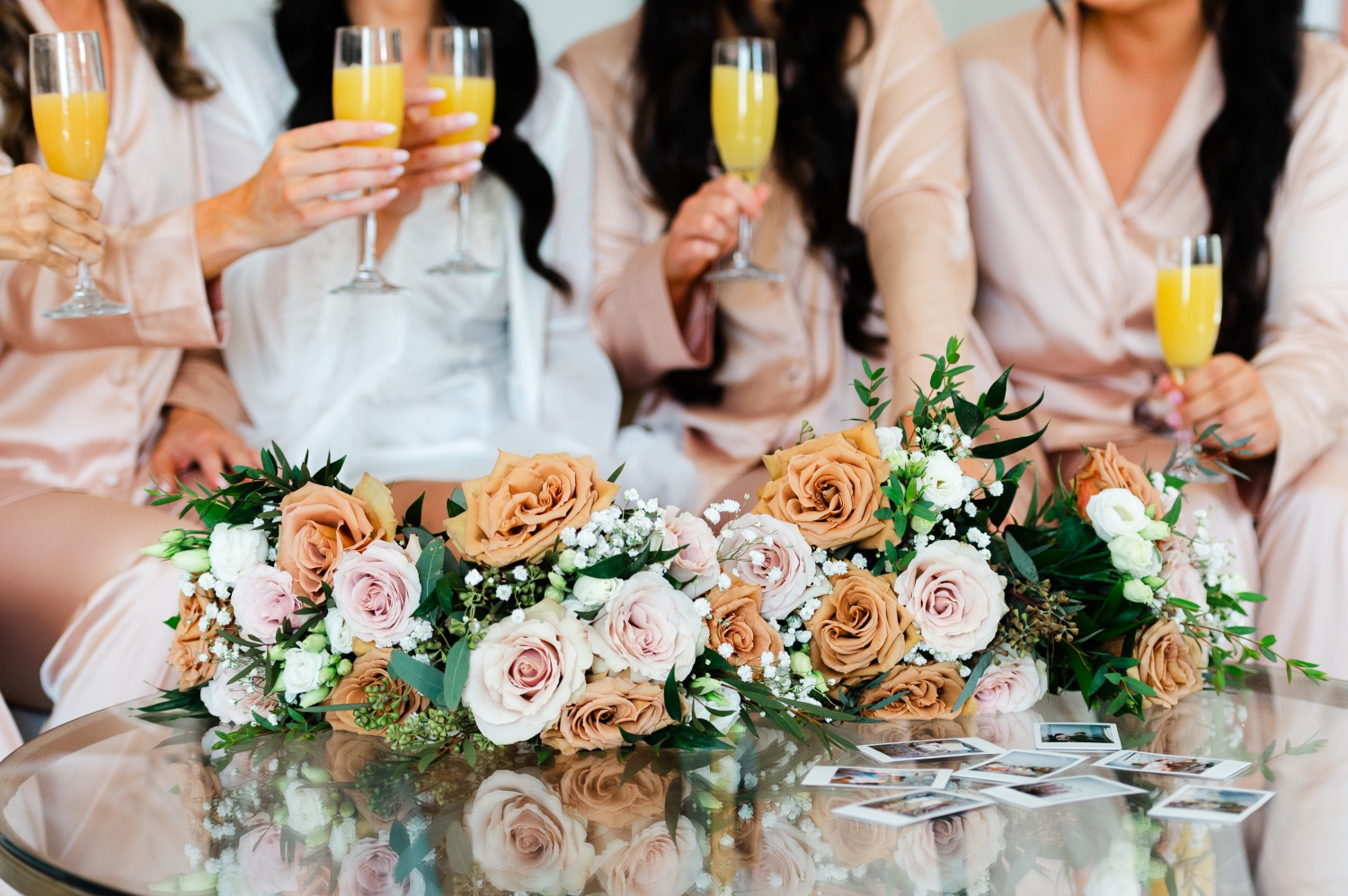 a photo of sand, peach and white wedding bouquets laying on a glass table with bridesmaids drinking orange juice champagne in the background. Wedding bouquets created by Ottawa wedding florist Acanthus Florals and photographed by JEMMAN Photography