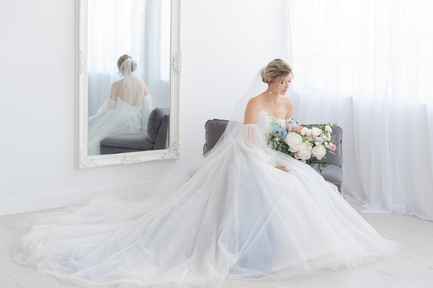 an elegant portrait of a bride sitting on a grey lounge chair holding a gorgeous bouquet of white peonies and blue florals created by Ottawa wedding florist The Gathering Event Co. Captured by JEMMAN Photography