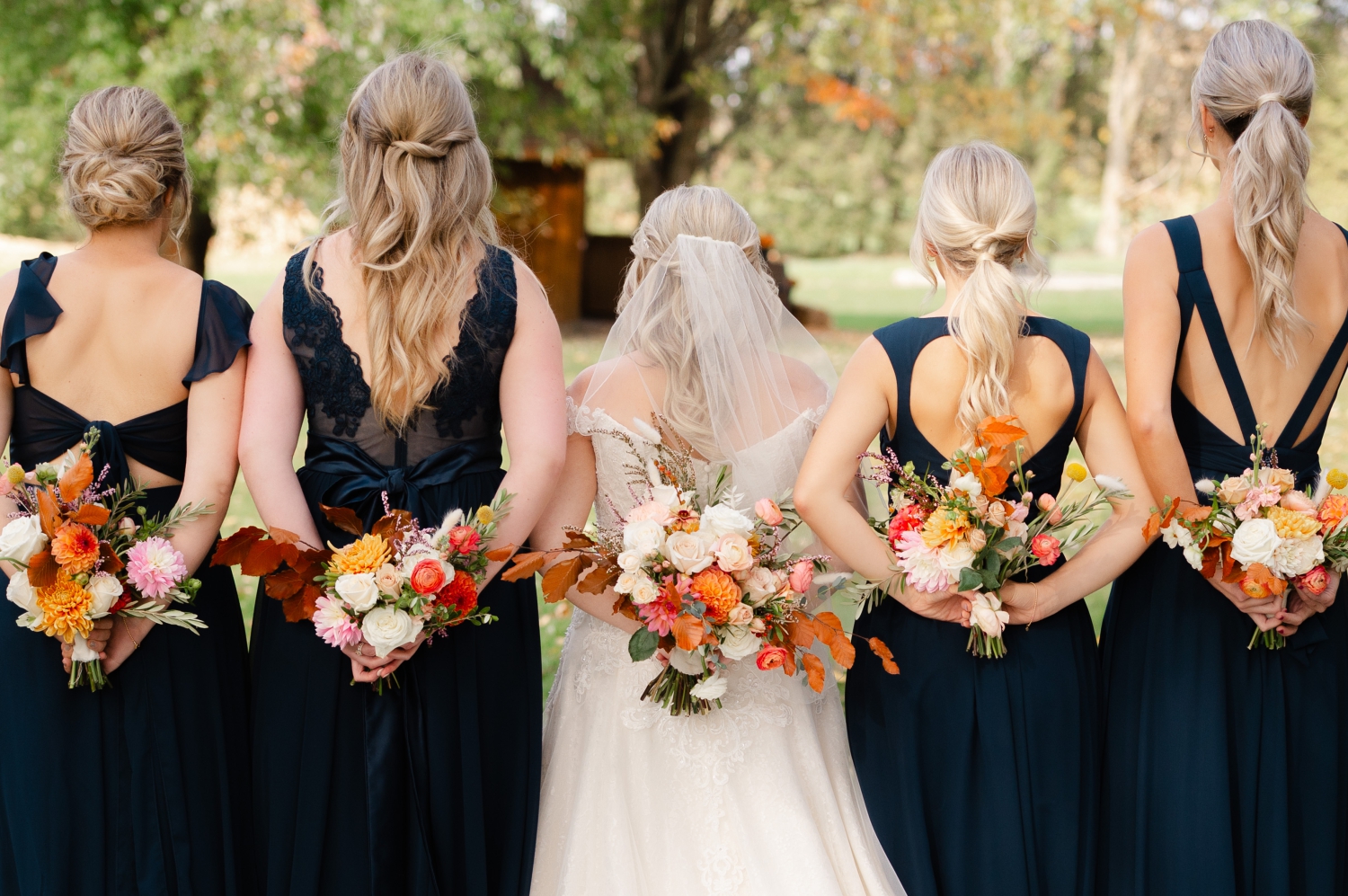 a bride and bridesmaids in blue dresses holding orange coloured wedding bouquets behind their backs to show off the back of their dresses. Wedding florals created by Riverwood Gardens and captured by JEMMAN Photography