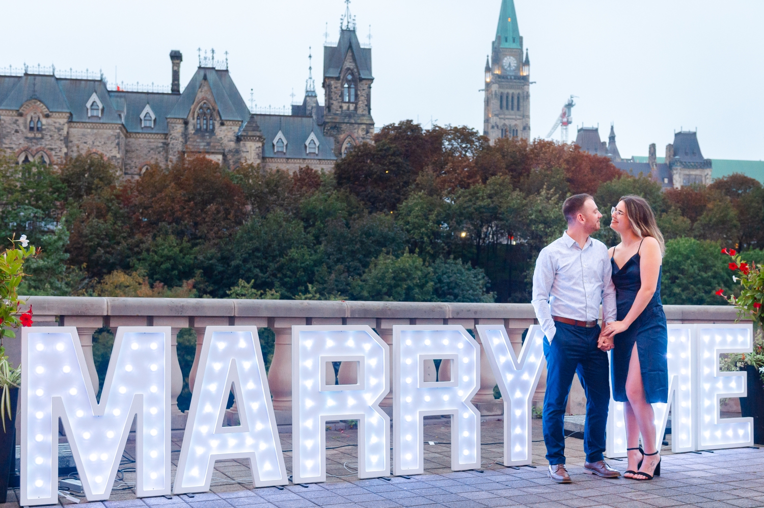 a male in a grey shirt and a female in a blue dress in front of a "Marry Me" sign with the Parliament buildings in the background as part of their surprise proposal in Ottawa. Captured by JEMMAN Photography