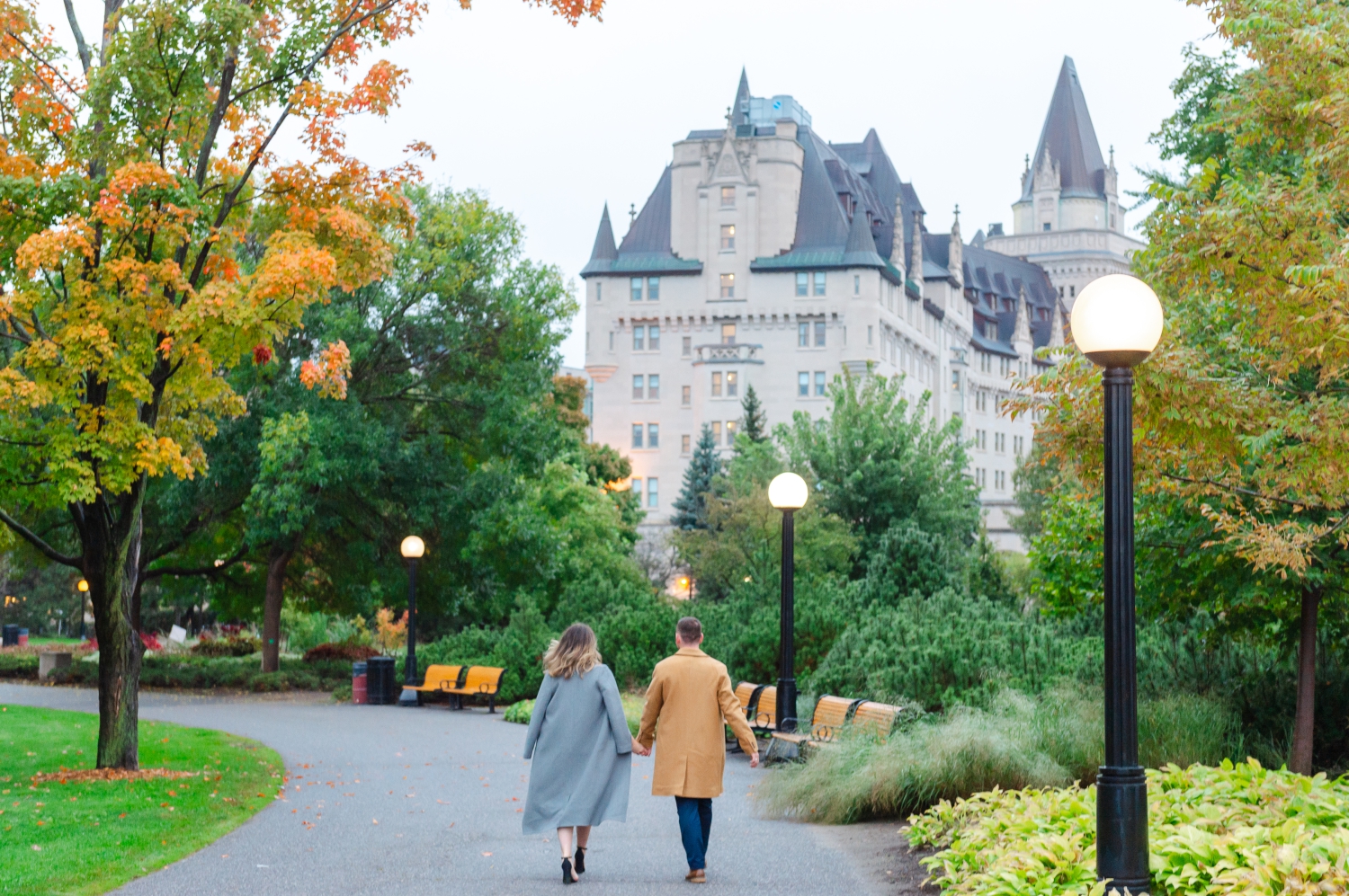 a male in a camel coloured jacket walking hand in hand with a female in a grey jacket along the paths of Major's Hill Park with the Chateau Laurier hotel in the background. Captured in Ottawa by JEMMAN Photography