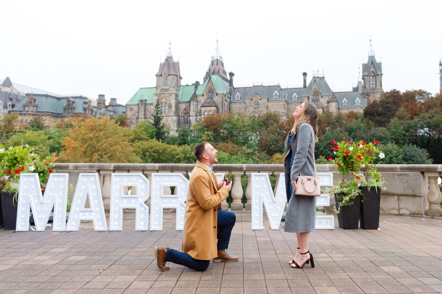 a photo of a male in a camel jacket on his knee proposing to a female in a grey jacket with the Parliament buildings in the background as part of his surprise proposal idea in Ottawa. Captured outdoors by JEMMAN Photography