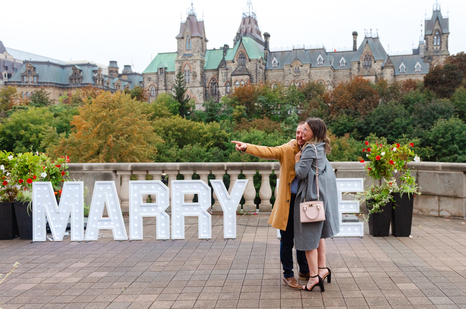 a male in a camel jacket pointing to his friends after he just proposed to his girlfriend in front of the Ottawa parliament buildings