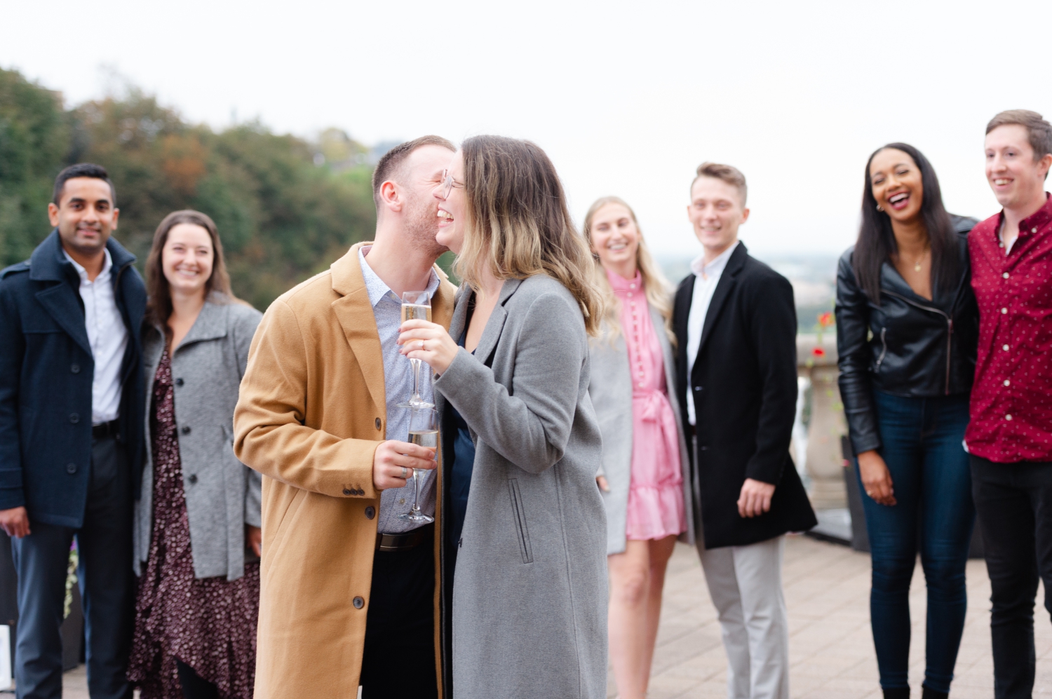 a male and female drinking champagne and kissing while their friends look on and smile after their surprise engagement proposal in Ottawa. Captured by JEMMAN Photography