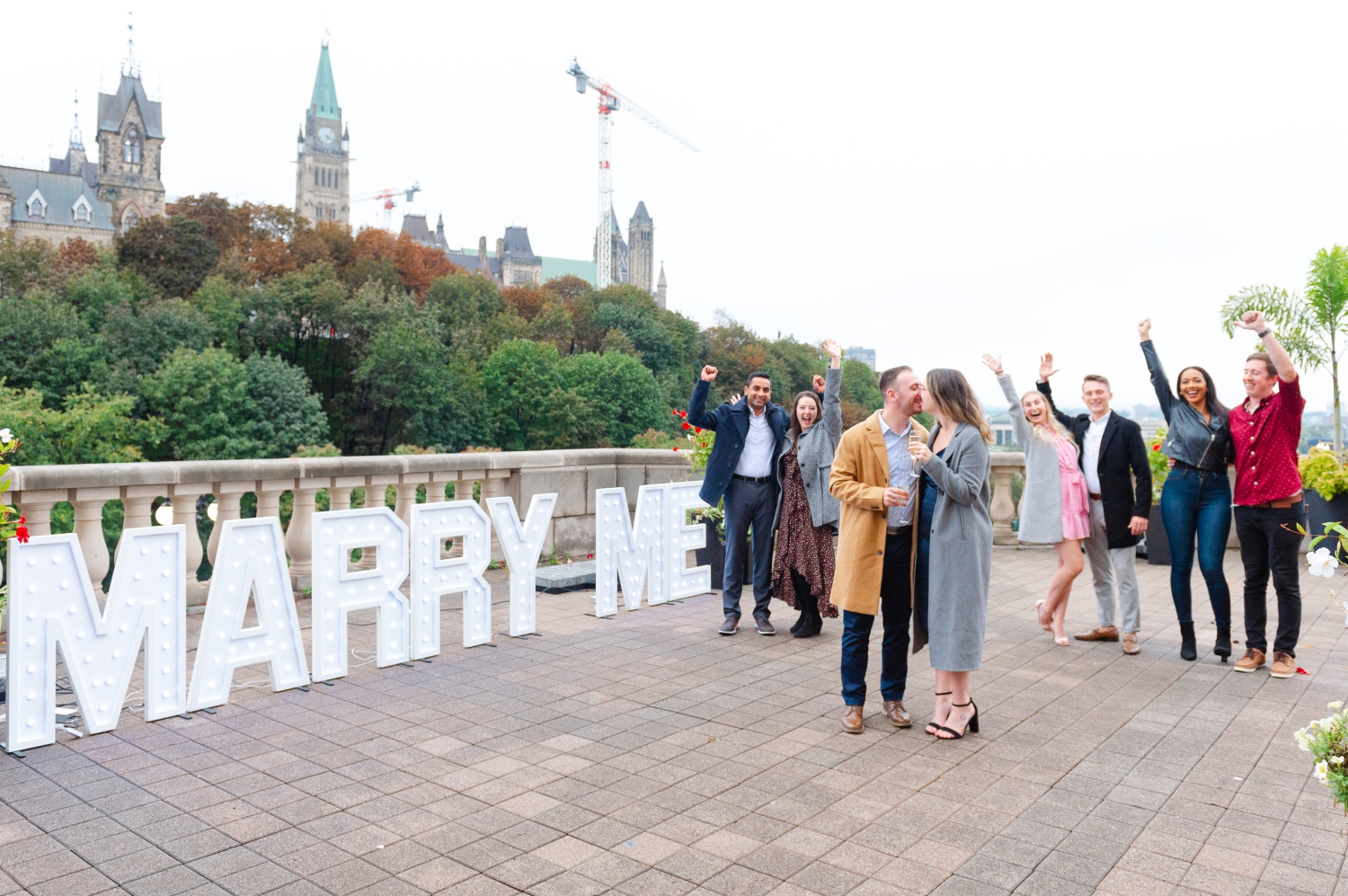 a male and female drinking champagne and kissing in front of a Marry Me sign while their friends look on and smile after their surprise engagement proposal in Ottawa. Captured by JEMMAN Photography