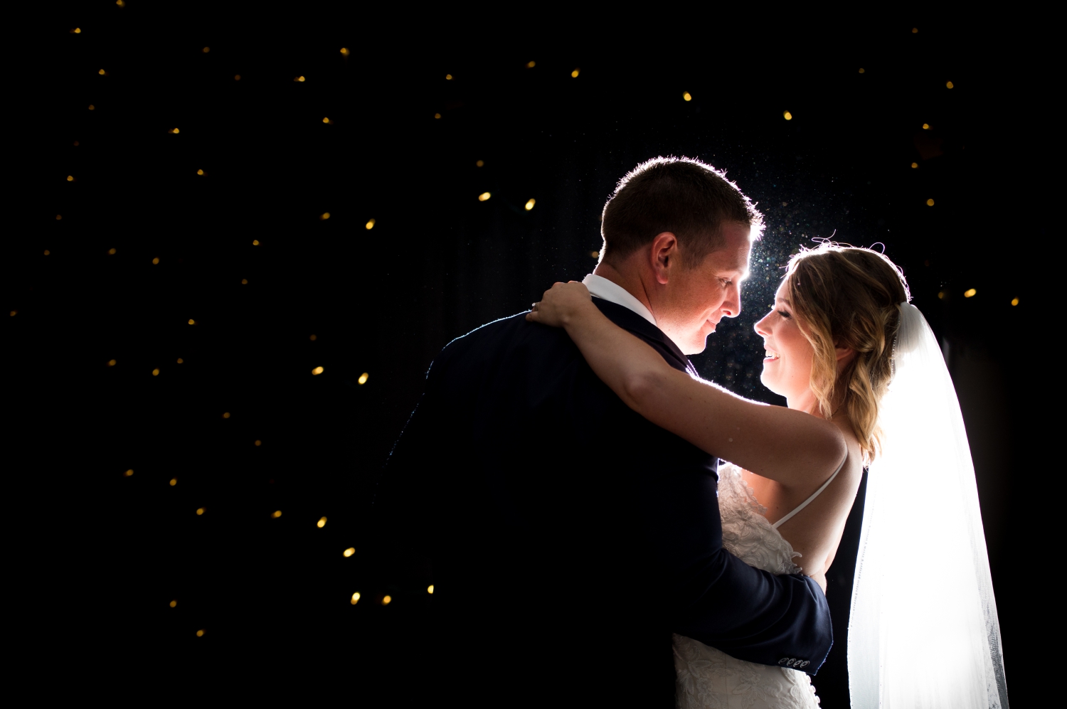 a backlit photo of a bride in a lace dress and veil smiling at her groom in a blue suit as they go through the steps of their wedding dance lessons in Ottawa