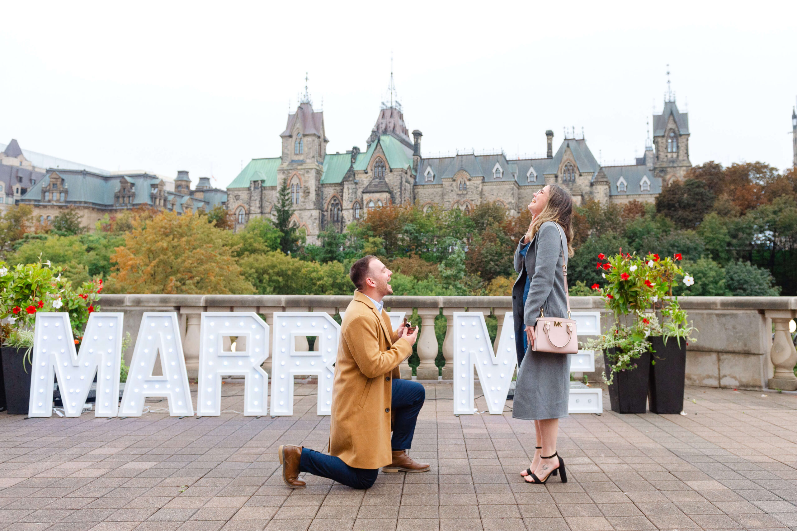 a male on one knee proposing to a female with a marry me sign and Parliament in the background - one of the best places to propose in Ottawa