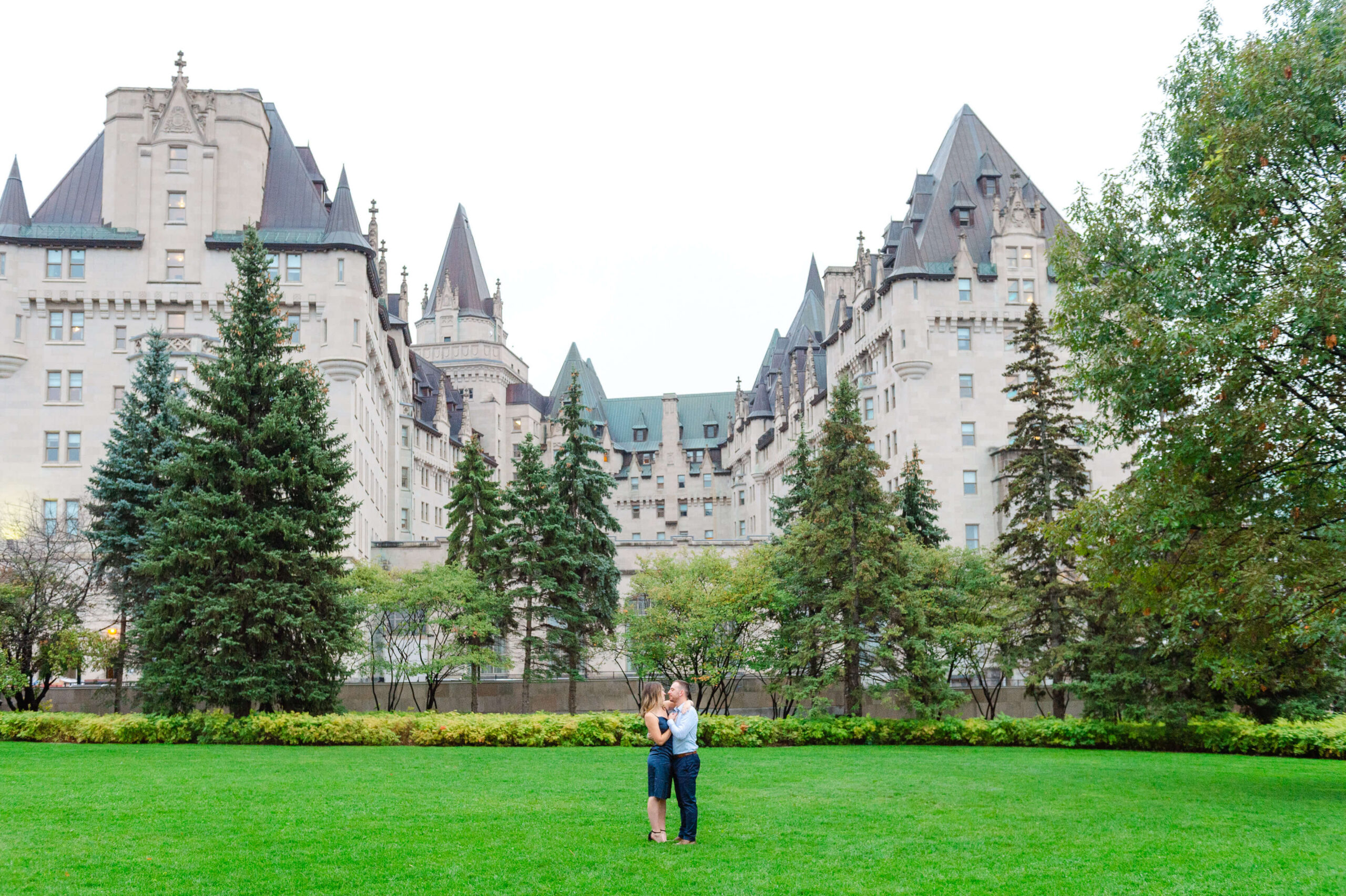 a male and female dressed in blue, kissing behind the Chateau Laurier, in Major's Hill Park, where they just got engaged. Captured by JEMMAN Photography