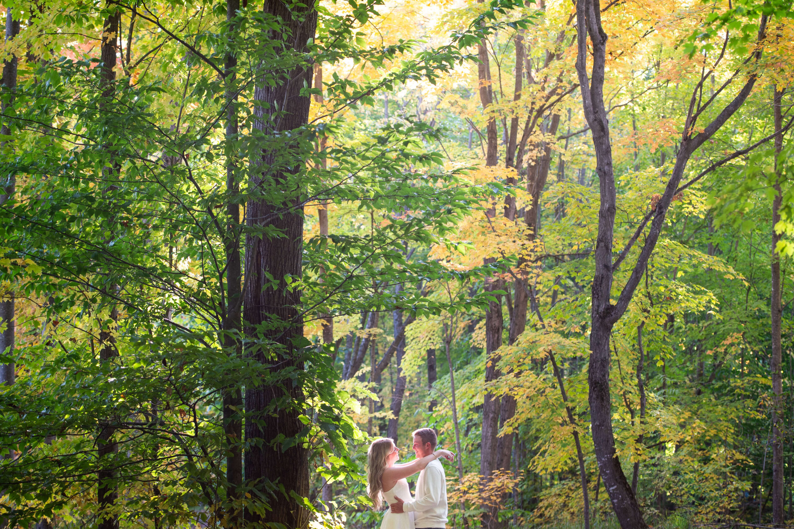 a male and female dressed in white smiling at each other surrounded by trees and the Gatineau forest, one of the best places to propose in Ottawa. Captured by JEMMAN Photography